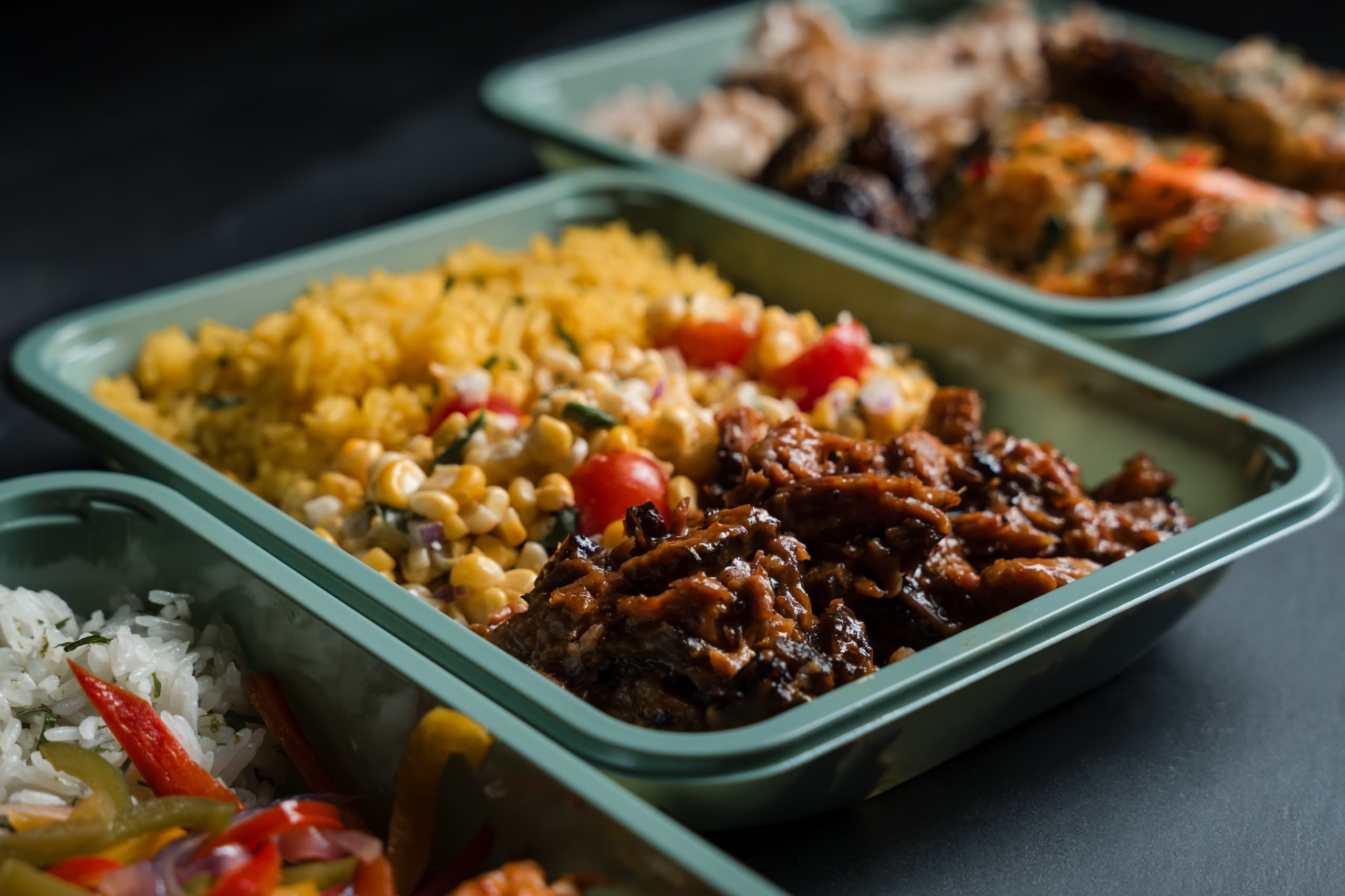 A line of sage-green ABLTY trays holding pulled meat, corn salad, and saffron rice