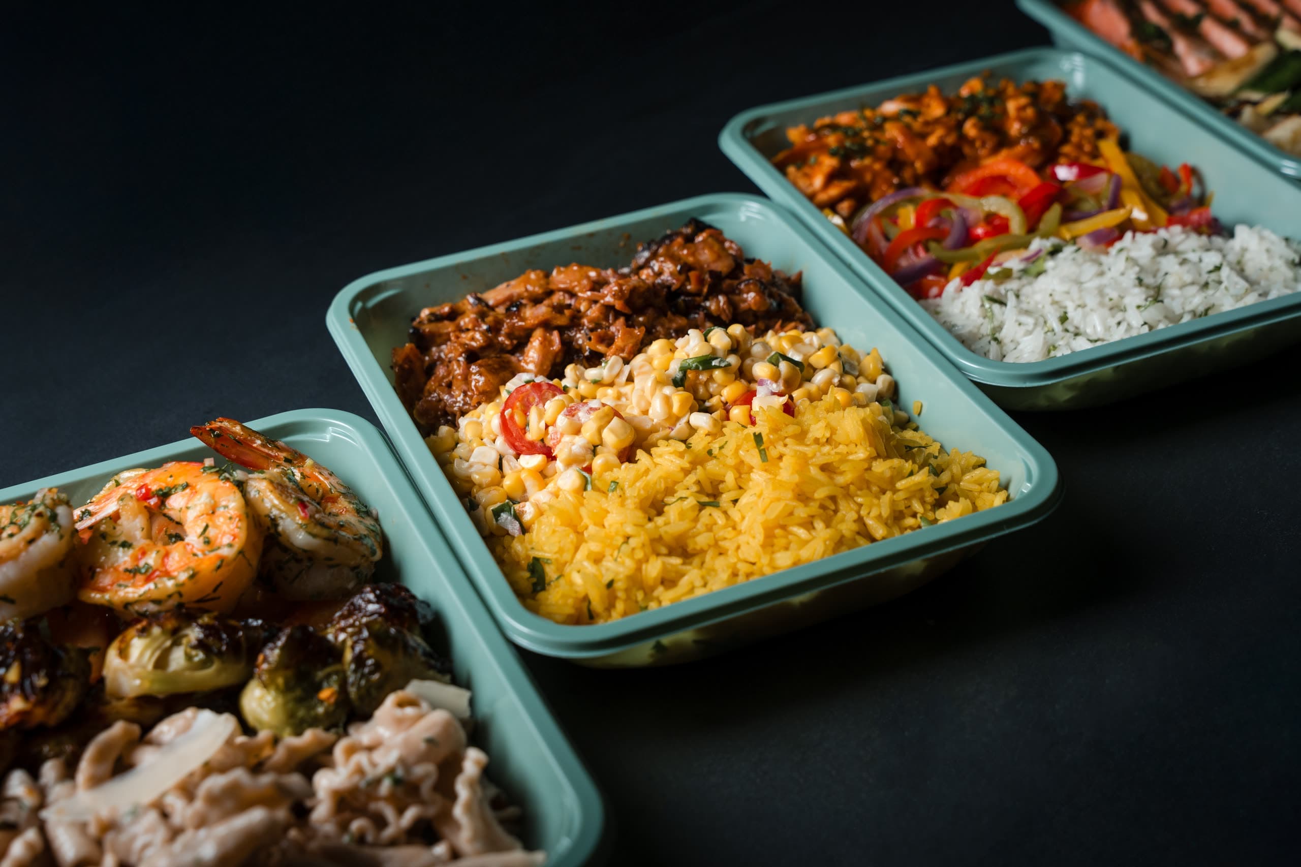 Three sage-green ABLTY meal trays receding on a diagonal — shrimp and brussels sprouts, glazed pork with corn salad and yellow rice, then a chopped salad with feta