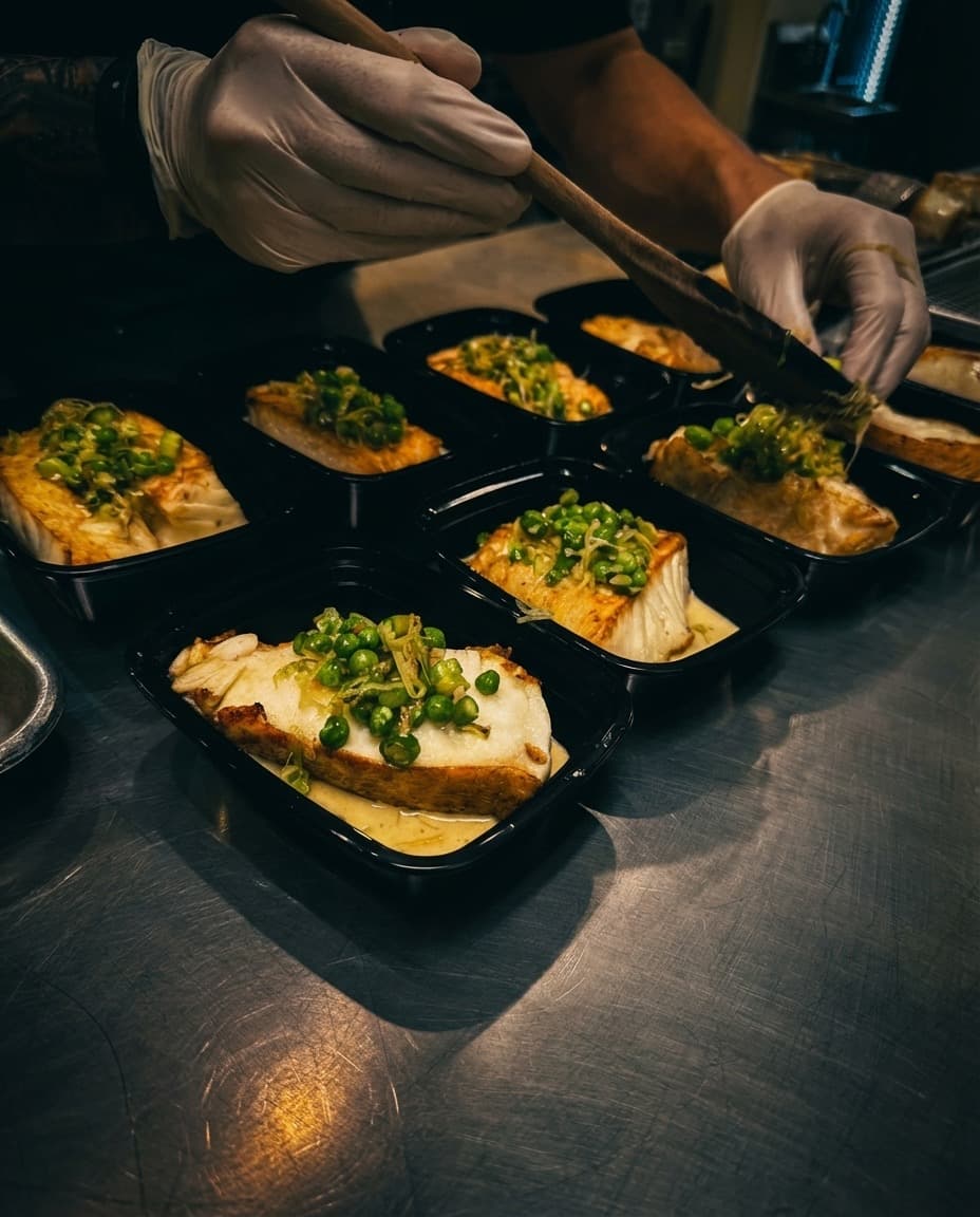 Gloved hands garnishing fish containers with peas on a stainless steel prep surface