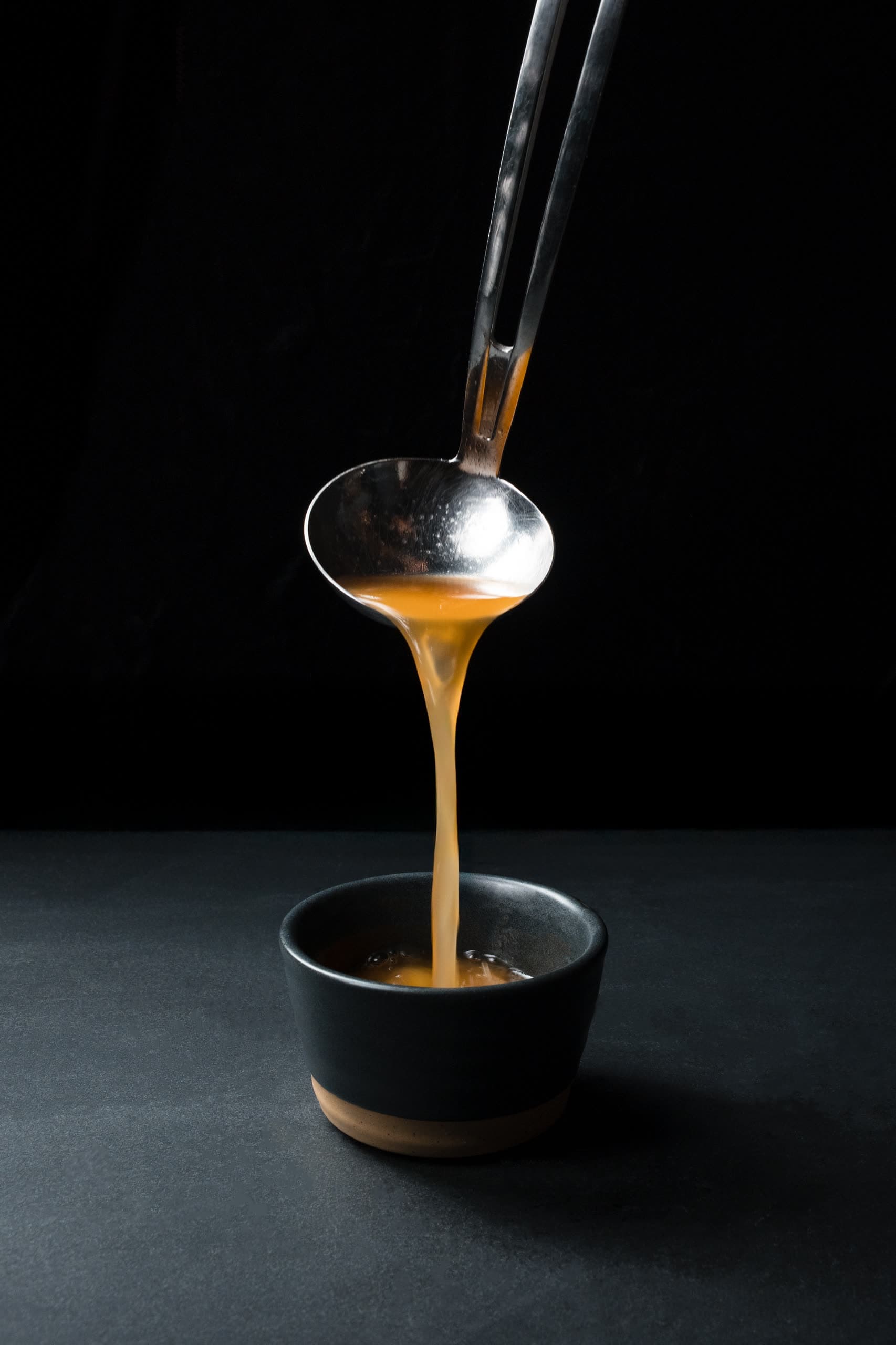 A steel ladle pouring golden broth into a small dark ceramic cup in the ABLTY kitchen