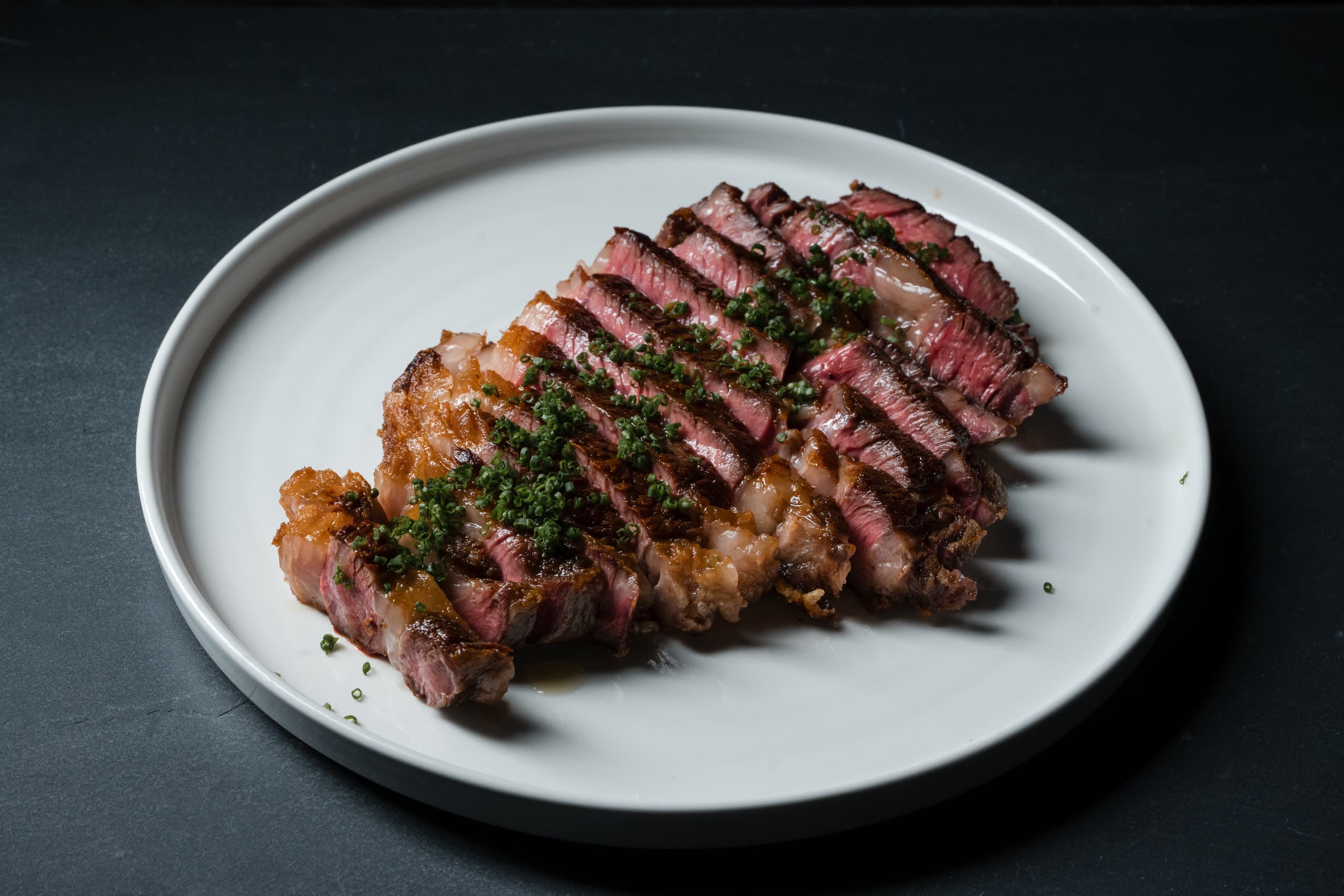 A sliced ribeye finished with chives on a white plate, rendered fat cap along one edge