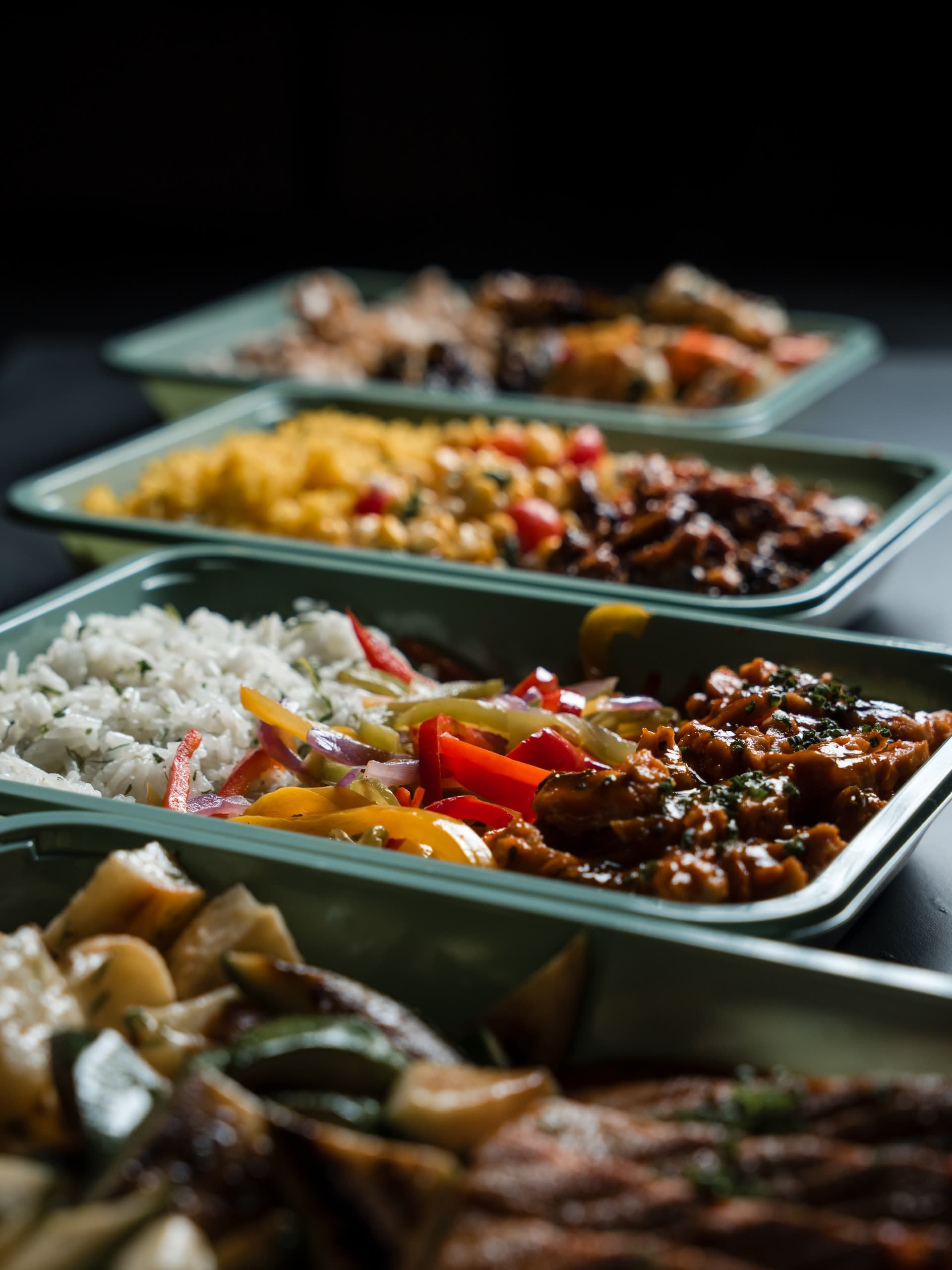 A receding line of sage-green ABLTY meal trays — herbed white rice, sautéed peppers and onions, and saucy braised chicken in the sharp tray, corn salad and yellow rice behind it