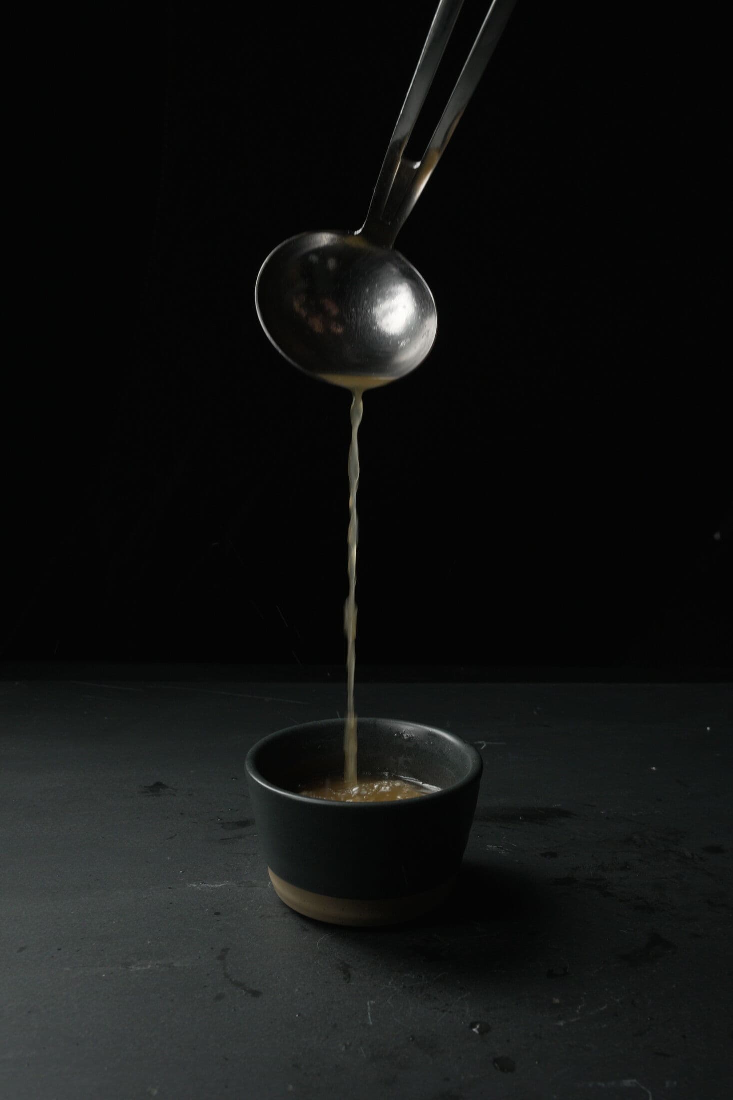 A steel ladle pouring golden broth into a small dark ceramic cup in the ABLTY kitchen