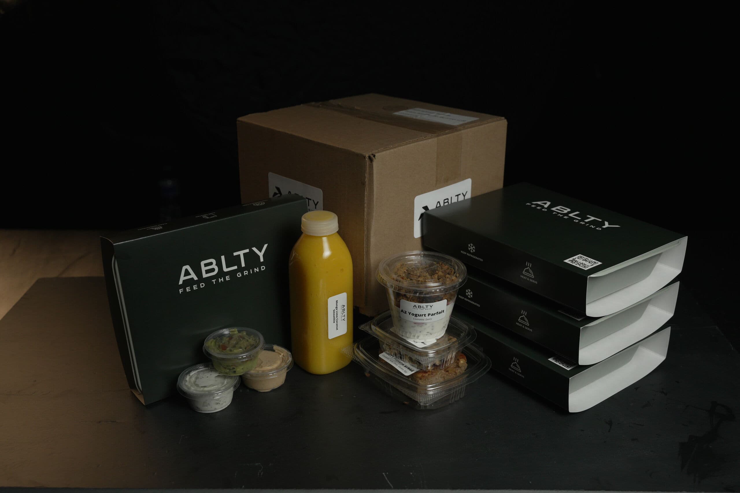 A packed ABLTY order — kraft shipping box, stacked heat-and-serve meal sleeves, a smoothie bottle, a yogurt parfait and sauce cups