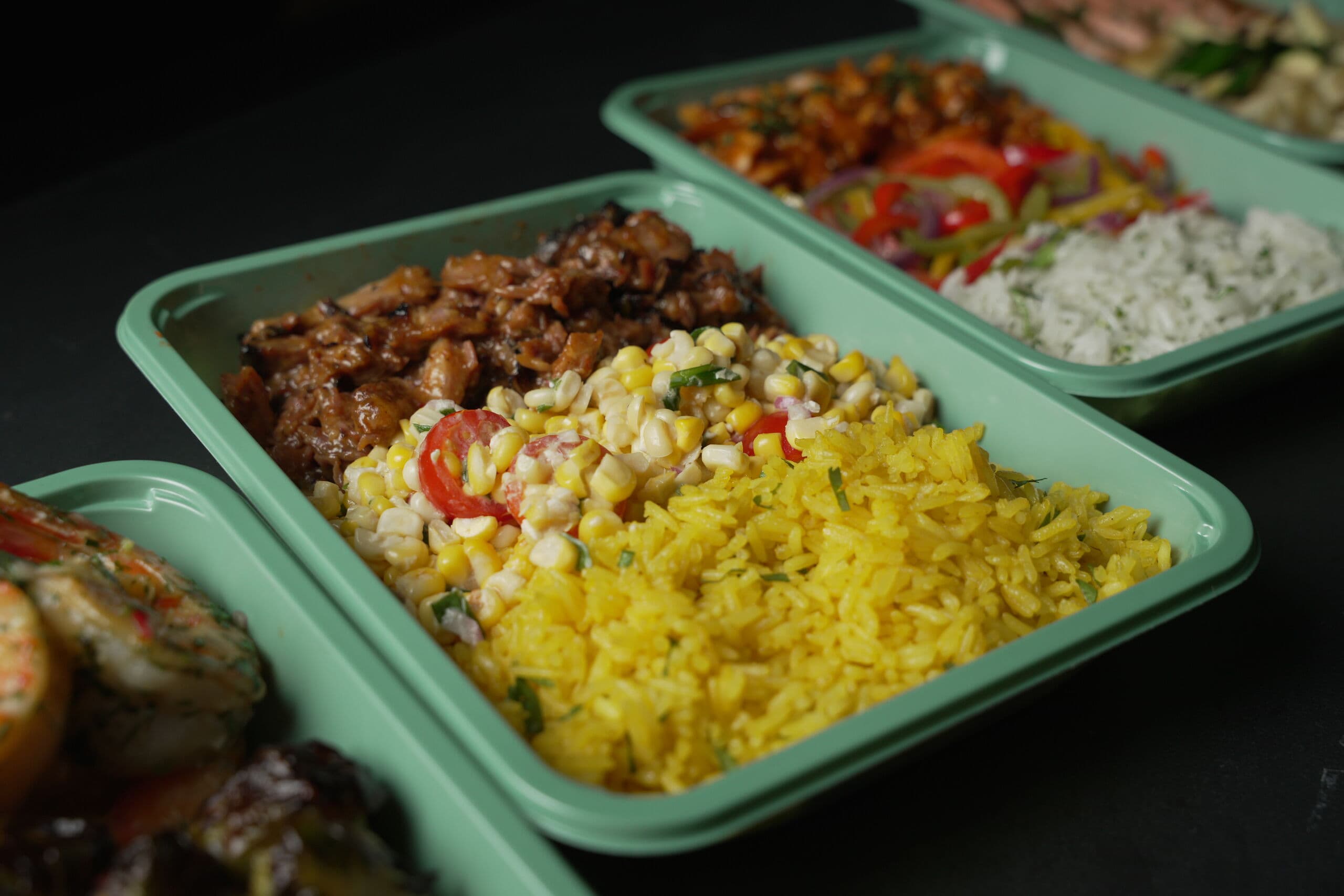 A line of chef-made ABLTY meal trays — glazed pulled meat with corn salad and yellow rice in front, shrimp, peppers, and salmon trays behind